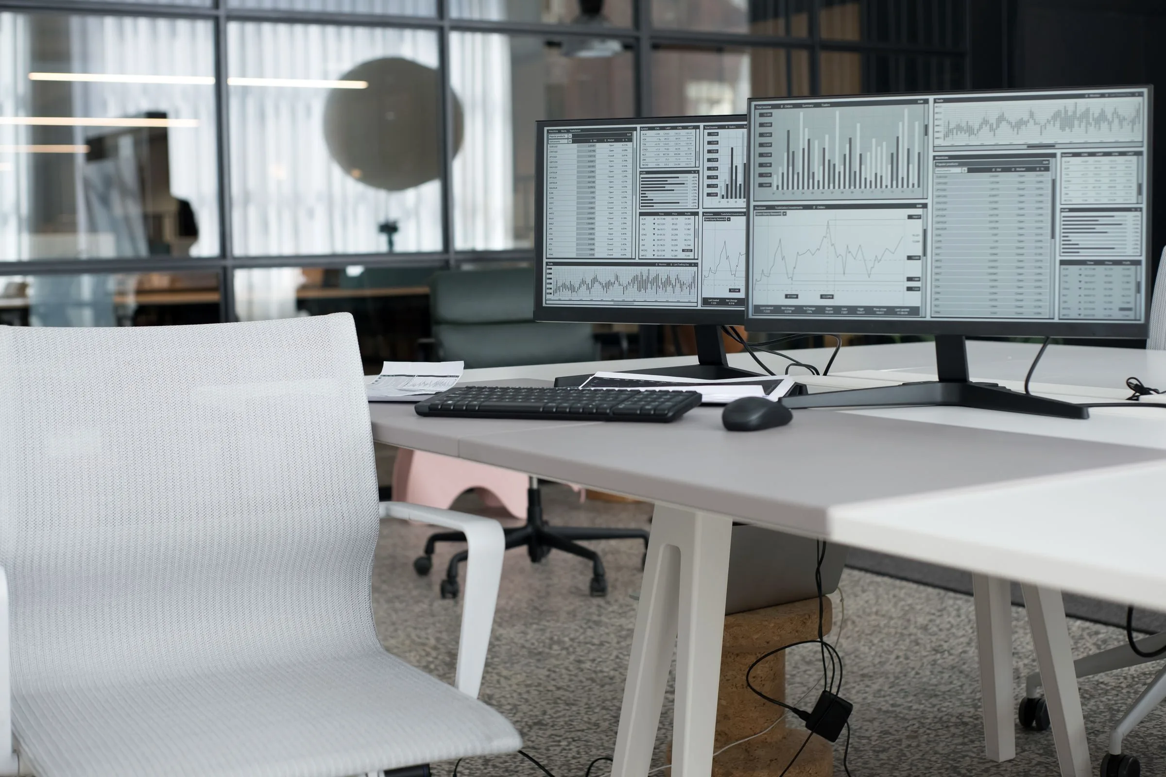 An office desk with dual monitors showing data charts, an ergonomic chair beside it.