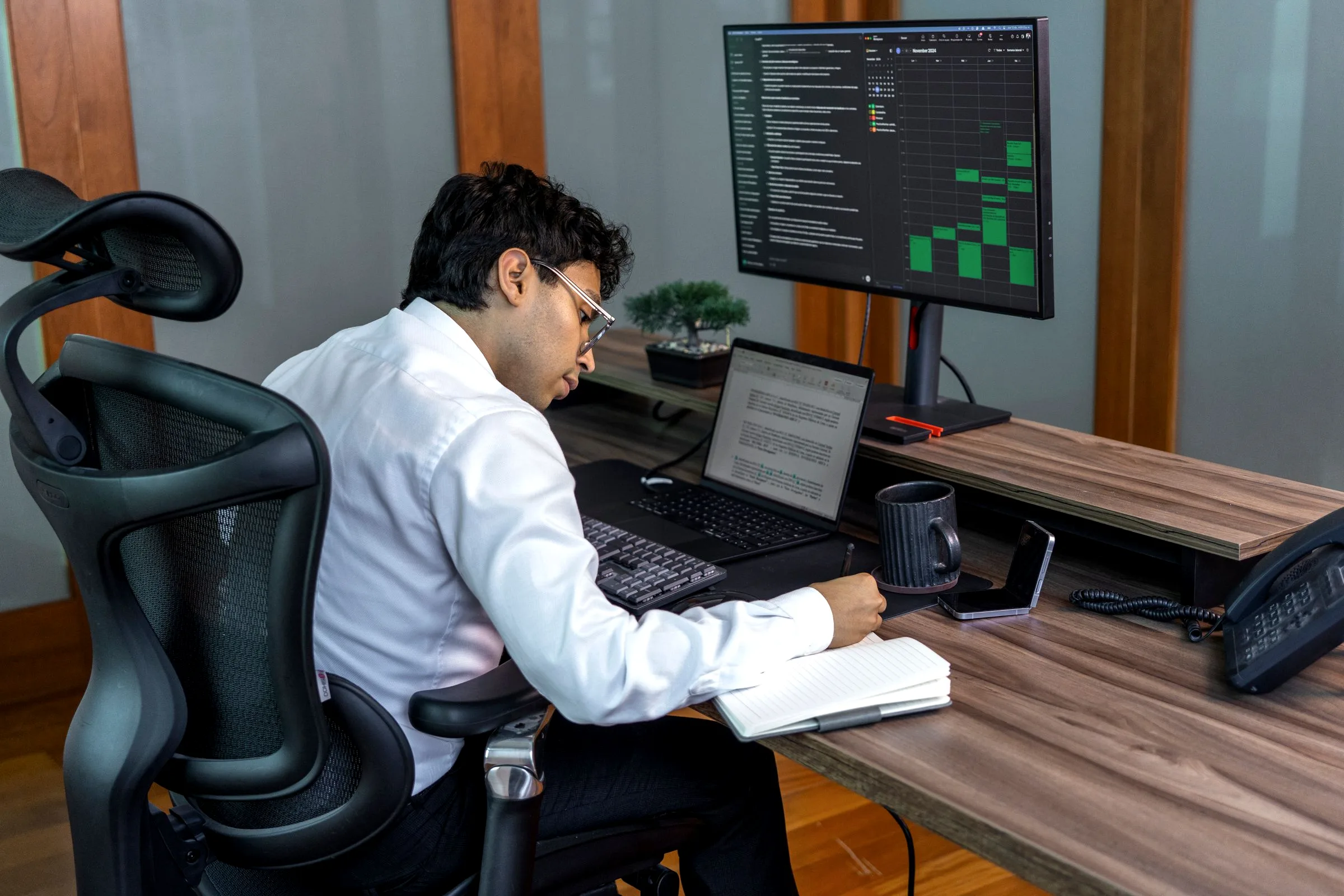 A man seen from behind at a desk, working at a laptop below a monitor showing a chart.