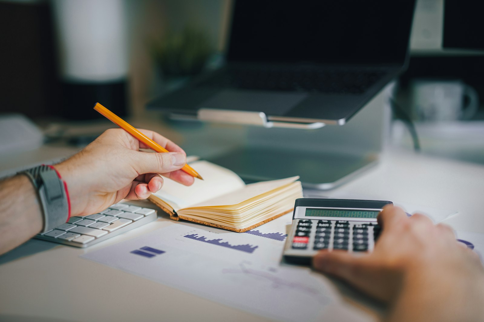 Close-up of hands working with a notebook and desk calculator beside a laptop